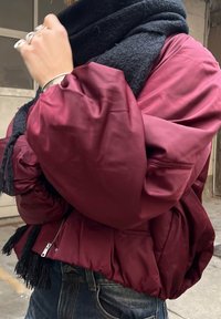 Burgundy puffer jacket with a shiny finish, featuring voluminous sleeves, side pockets, and a zip closure, paired with a textured black scarf.