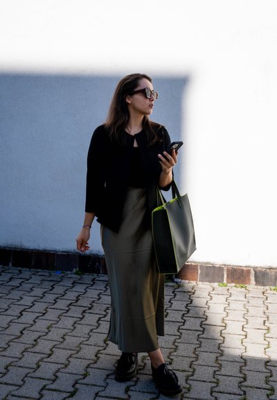 Chaqueta negra, falda verde oliva, gran bolso negro con acentos verdes brillantes, gafas de sol, sosteniendo un teléfono inteligente, de pie sobre una superficie pavimentada.