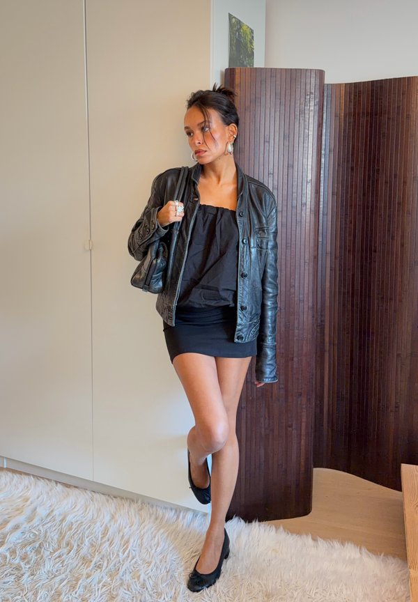 Mujer con chaqueta de cuero negra, minifalda negra y zapatos negros, de pie en el interior frente a una pared blanca y panel de madera oscura.