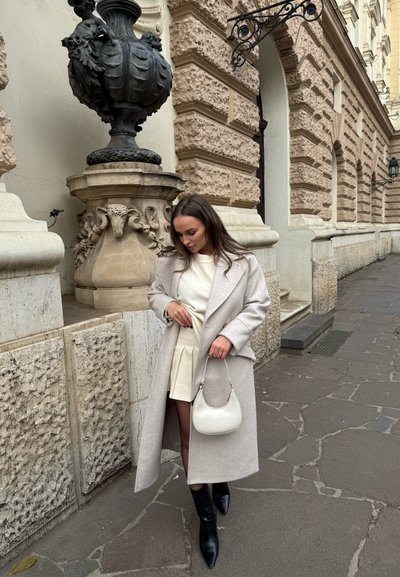 Abrigo largo gris claro sobre un suéter blanco, combinado con una falda mini color crema y botas negras hasta la rodilla. Sosteniendo un pequeño bolso blanco.