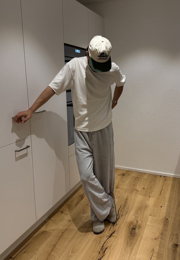 Person trägt weißes Hemd, weite graue Hose und Mütze und lehnt an einem Küchenschrank, auf dem ein Holzboden zu sehen ist.