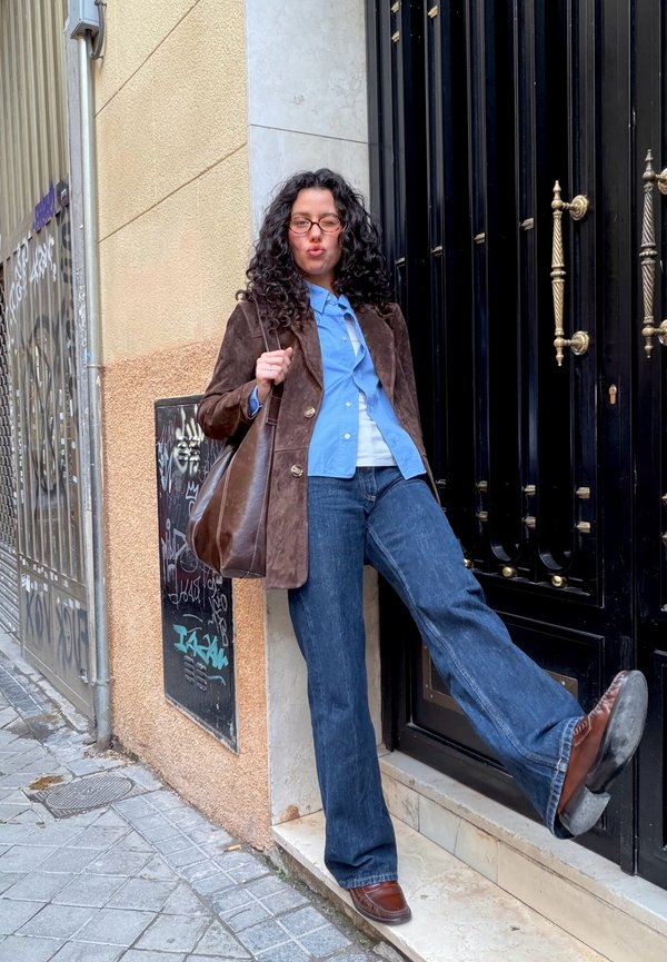 Personne aux cheveux bouclés, portant des lunettes, une veste marron, une chemise bleue, un jean et des chaussures marron, appuyée contre une porte noire, levant une jambe sur une marche.