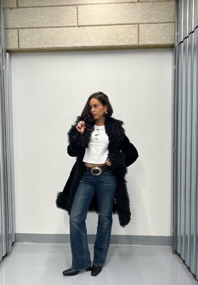 Mujer posando con un abrigo de piel sintética negra, top blanco corto, vaqueros azules acampanados, botas negras y sosteniendo gafas de sol cerca de sus labios.