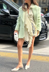 Manteau en vert menthe, duveteux, sur une chemise crème avec des volants, associé à un détail latéral pailleté, un sac à main blanc et des chaussures à petit talon beige.