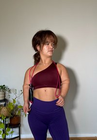 Top sportivo bordeaux con una spallina, abbinato a leggings viola a vita alta con texture. Una corda per saltare rosa pende dal reggiseno.