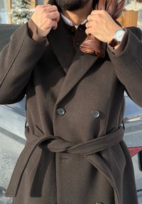 pardessus brun croisé avec ceinture, en tissu épais. Il est orné de grands boutons noirs et possède un col montant. Des gants en cuir brun sont tenus à la main.