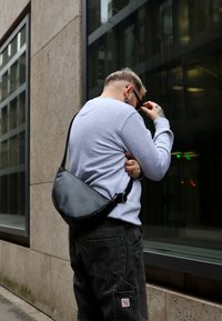 Fekete bőrből készült keresztpántos táska ívelt formával, állítható pánttal és sima felülettel, egy világosszürke pulóver és sötét nadrág fölött viselve.