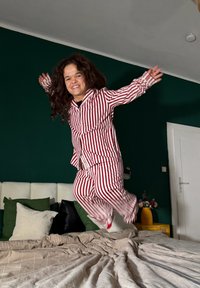 Personne en pyjama rayé rouge et blanc sautant sur un lit avec des oreillers verts et blancs dans une chambre vert foncé.