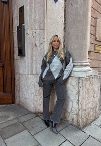 Jeune femme aux longs cheveux blonds portant un pull gris à motif losanges, un jean noir et des bottes noires pointues, appuyée contre un coin de bâtiment en marbre.