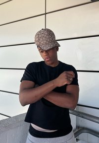 Black short-sleeved shirt, grey trousers, brown patterned cap. Well-built, arms crossed, standing against a minimalist background.