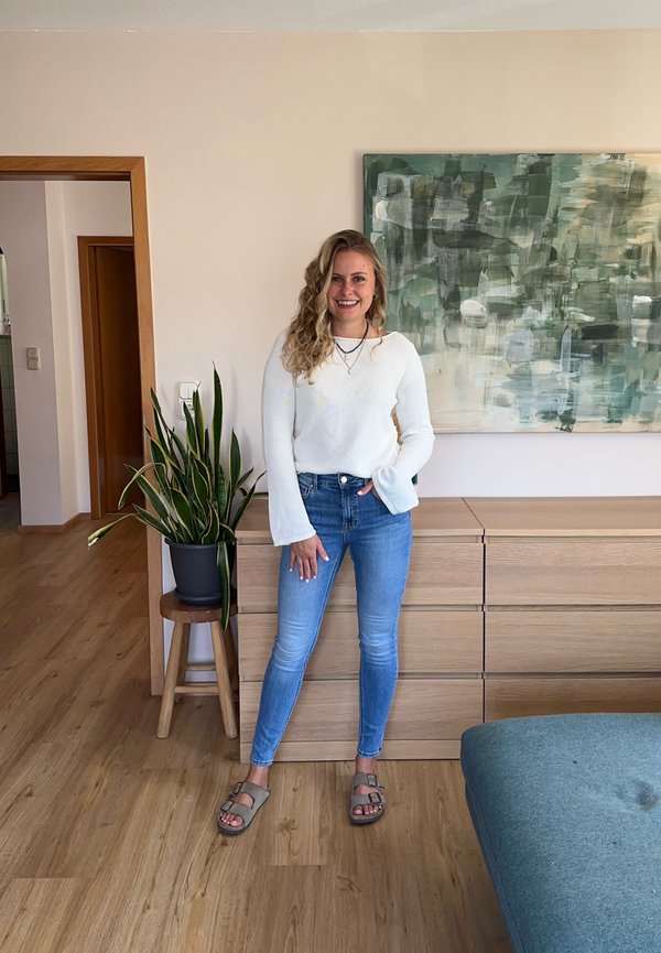 Vrouw met krullend haar in een witte trui en spijkerbroek staat glimlachend bij een houten kastje en een groene plant in een moderne woonkamer.