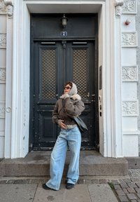 Jeune personne portant un jean trop grand, une veste marron et un sweat à capuche, debout devant une grande porte double noire avec un encadrement blanc orné.