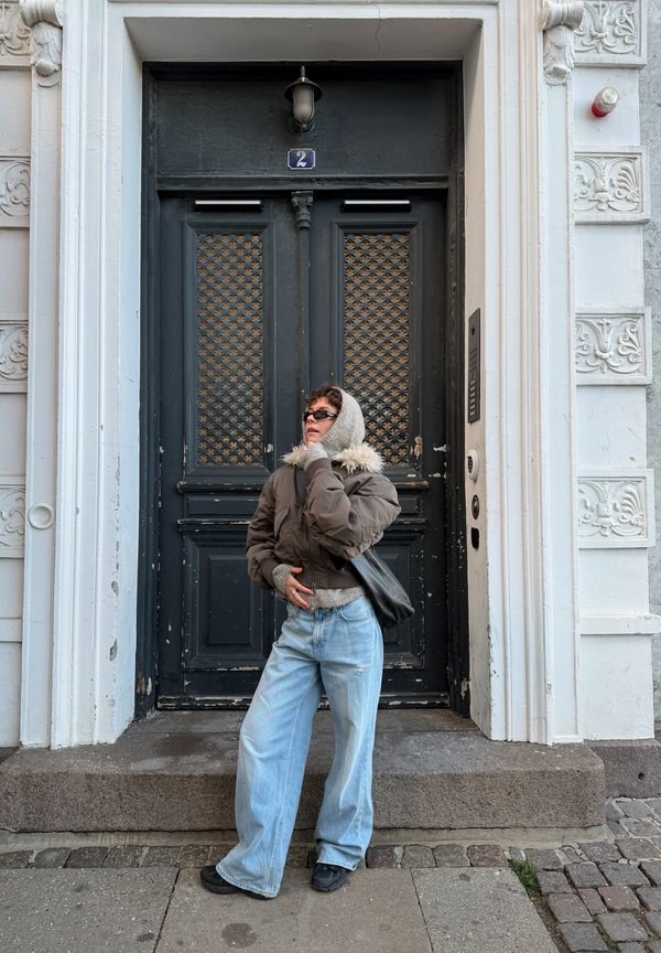 Young person dressed in oversized jeans, a brown jacket, and a hooded jumper stands against a large black double door with ornate white framing.