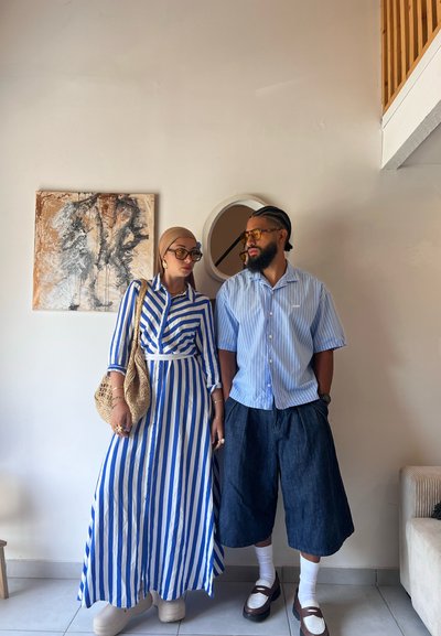 Dos personas están juntas en el interior. La mujer lleva un vestido largo a rayas azul y blanco y un pañuelo en la cabeza, y sostiene un bolso de mimbre. El hombre viste una camisa de manga corta a rayas azul clara y pantalones cortos de mezclilla oscuros, con calcetines blancos y zapatos marrones.