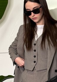 Femme aux longs cheveux bruns portant des lunettes de soleil noires et un costume gris trois pièces, tenant une pochette noire Givenchy sur un fond blanc.