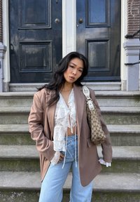 Brun oversized blazer over en hvid blondebluse med flæser, parret med lyseblå højtaljede jeans og en beige mønstret skuldertaske.