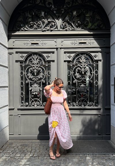 Mujer con vestido rosa estampado y chanclas se retoca el cabello, de pie sobre adoquines frente a grandes puertas dobles de metal gris ornamentadas.