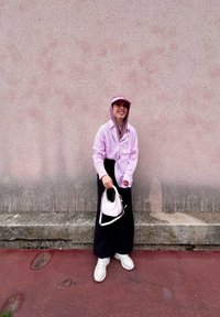 Ροζ πουκάμισο με κουμπιά, μαύρα παντελόνια σε φαρδιά γραμμή, λευκά αθλητικά παπούτσια και ροζ καπέλο. Κρατάει μια μικρή ροζ τσάντα χεριού με ιμάντα στον ώμο.