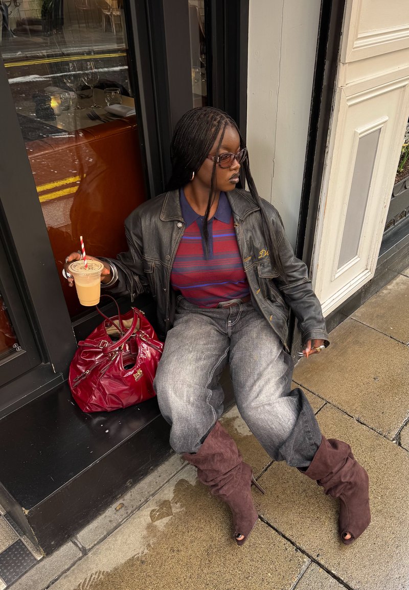 Žena s vrkočmi, ktorá má na sebe slnečné okuliare, koženú bundu, pruhované tričko a topánky, sedí pred kaviarňou, drží ľadovú kávu a cigaretu.