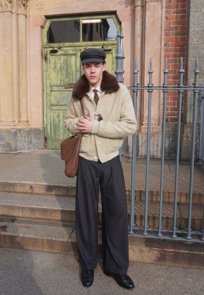 Joven de pie en unos escalones de piedra junto a una valla de hierro, vestido con una chaqueta crema con cuello de piel, pantalones anchos oscuros, gorra plana y un bolso marrón al hombro.