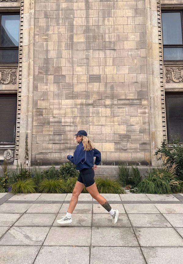 Una mujer con una gorra y una chaqueta azul marino, pantalones cortos negros y zapatillas blancas pasa junto a la fachada de piedra de un edificio.
