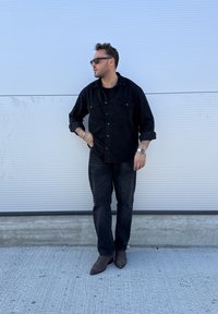 Čierna rifľová košeľa s náprsnými vreckami, čierne tričko, čierne rifle a hnedé špicaté topánky. Muž nosiaci slnečné okuliare stojí pred bielym pozadím.