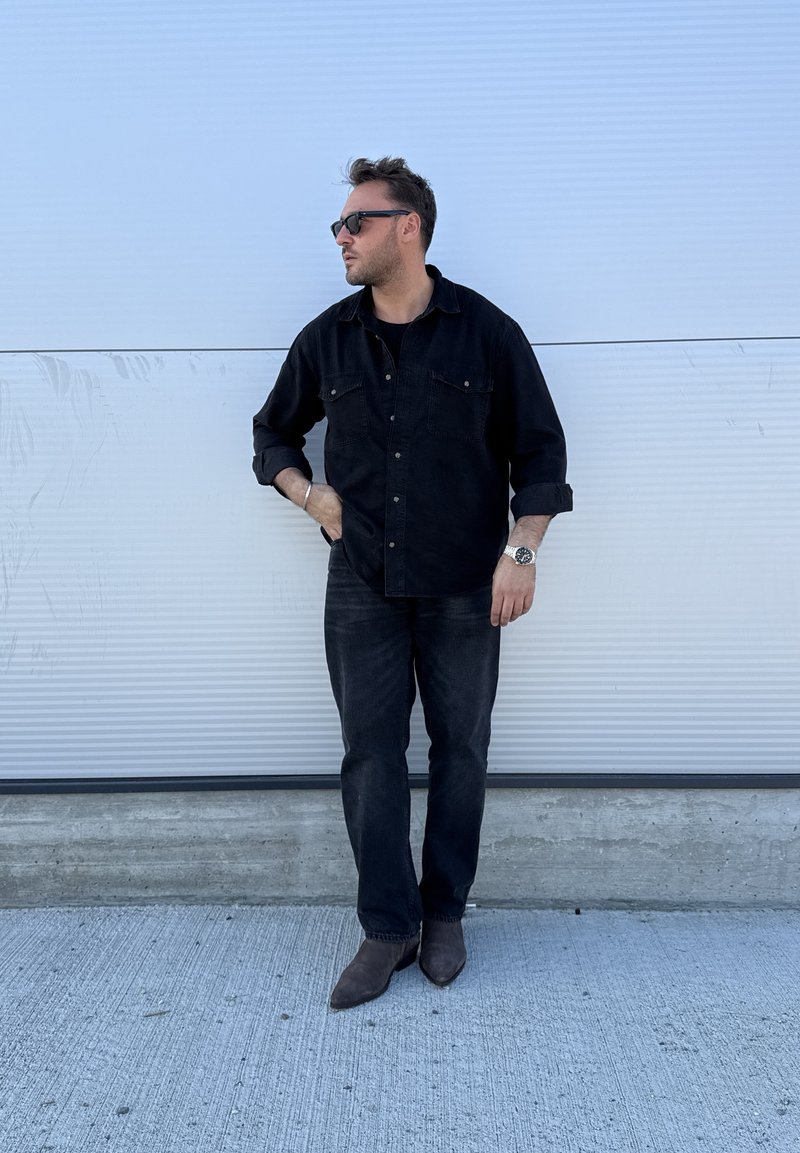 Schwarzes Denimhemd mit Brusttaschen, schwarzes T-Shirt, schwarze Jeans und braune spitze Stiefel. Mann mit Sonnenbrille, stehend vor weißem Hintergrund.