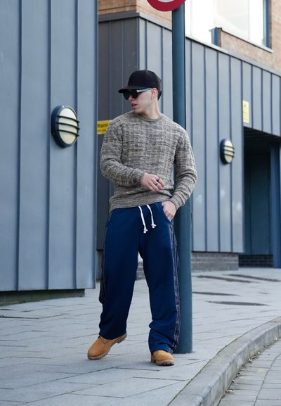 Suéter gris de punto, pantalones de chándal azul marino con rayas laterales y zapatos color beige. Los accesorios incluyen gafas de sol negras y una gorra negra. Entorno urbano.