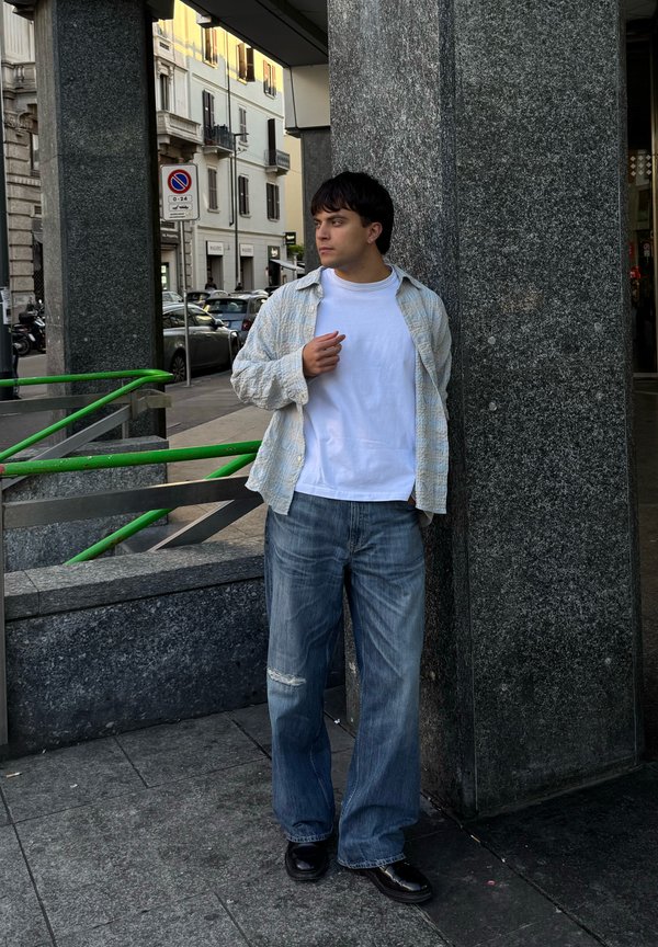 Leger Outfit mit einem hell karierten Hemd über einem weißen T-Shirt, kombiniert mit ausgewaschenen blauen Jeans und schwarzen Schuhen, an eine graue Wand gelehnt.