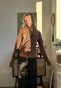 Jeune femme en sweat à capuche à carreaux et pantalon noir debout à l'intérieur près d'un meuble, la lumière du soleil projetant des ombres sur elle et dans la pièce.