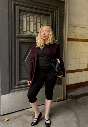 Blazer burdeos sobre una blusa negra, combinado con leggings capri negros y zapatos planos negros. El cabello rubio ondulado y un bolso negro completan el look.