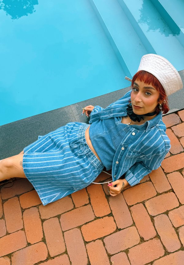Personne dans une tenue à rayures bleues et chapeau blanc, allongée sur des briques au bord d'une piscine, regarde la caméra avec une expression détendue.