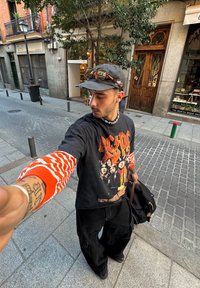 T-shirt preta com gráfico do AC/DC, manga longa listrada em vermelho e laranja por baixo, calças largas pretas, carregando uma bolsa castanha, ao ar livre.