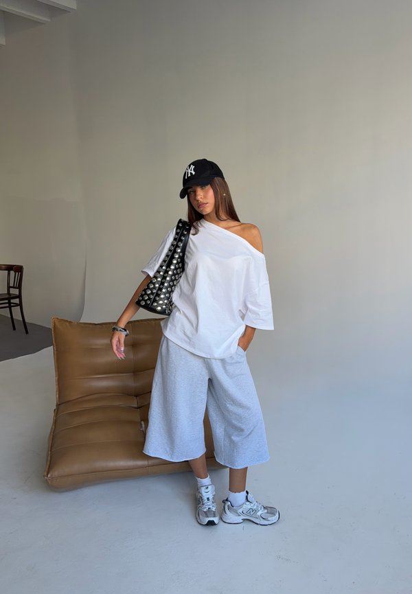 Kobieta w białym oversize t-shircie i szortach, czarnej czapce, białych trampkach i czarnej torbie z ćwiekami stoi obok brązowego tapicerowanego krzesła w minimalistycznym pokoju.