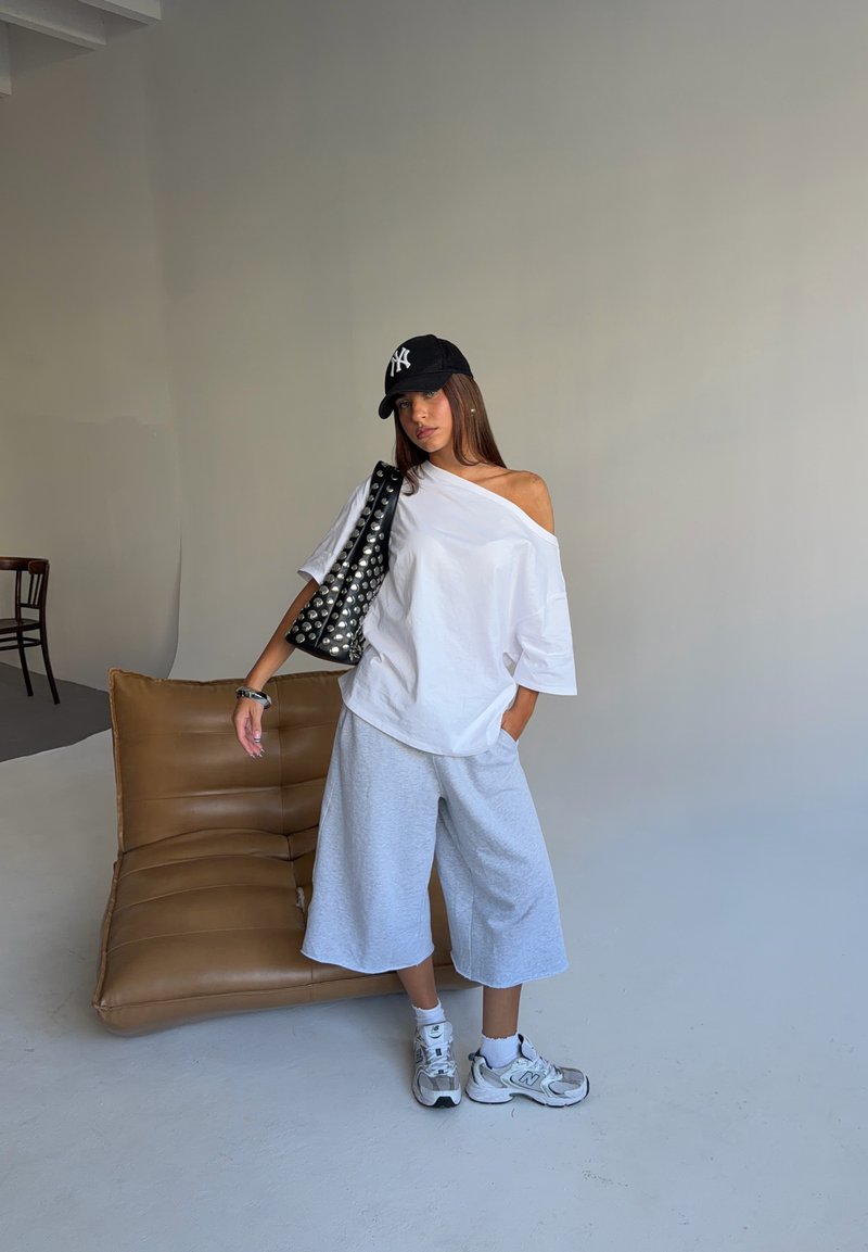 Γυναίκα με λευκό oversized μπλουζάκι και σορτς, μαύρο καπέλο, λευκά αθλητικά παπούτσια και μαύρη τσάντα με καρφιά στέκεται δίπλα σε μια καφέ μαξιλαρωμένη καρέκλα σε έναν μινιμαλιστικό χώρο.