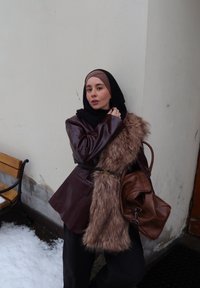 Jeune femme portant un manteau superposé en cuir marron et fausse fourrure avec un sac marron, debout près d'un banc couvert de neige en plein air.