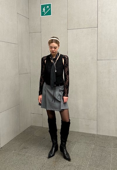 Blusa negra de manga larga con detalles de encaje, falda gris plisada, botas negras hasta la rodilla y una corbata negra con acentos plateados.