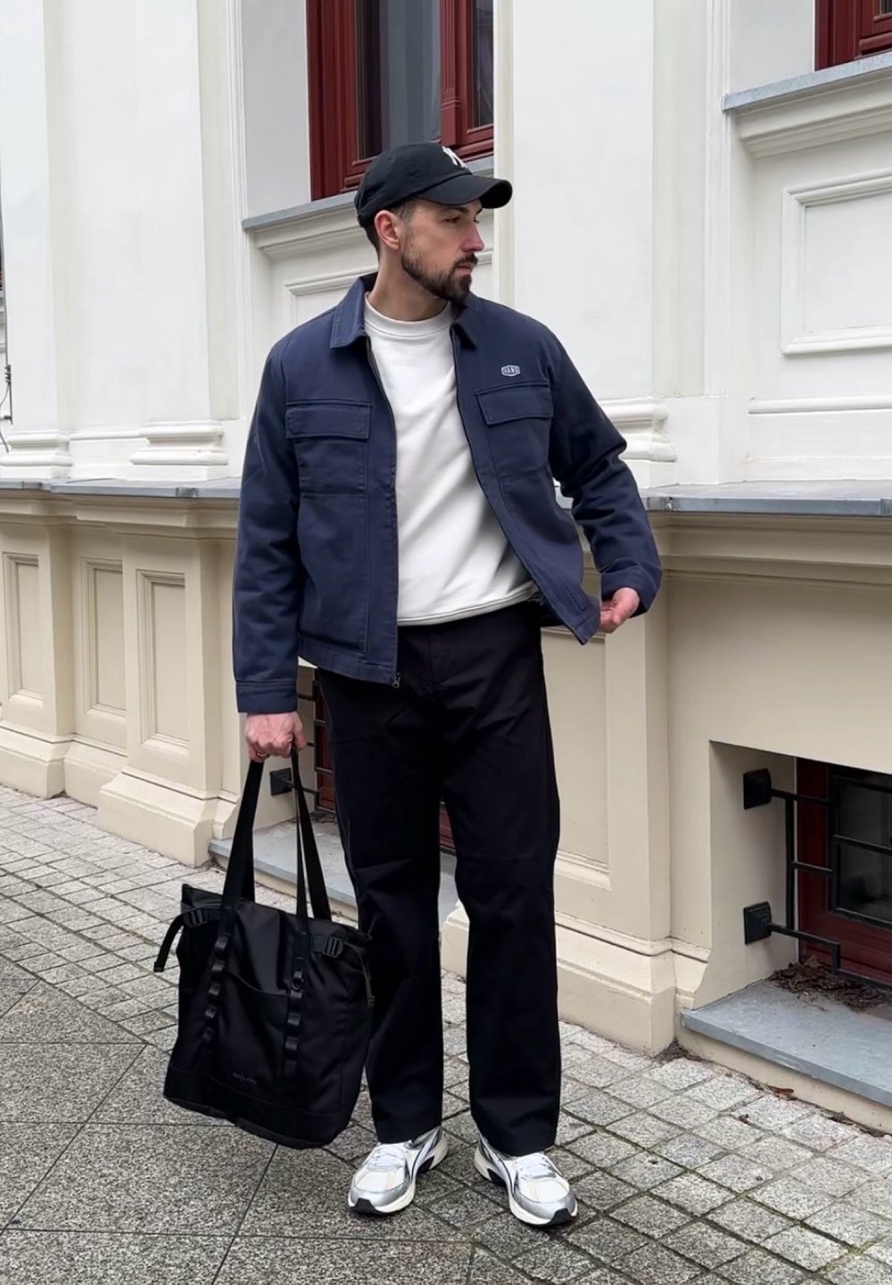 Veste bleu marine avec deux poches avant, sweat-shirt blanc, pantalon noir, portant un sac fourre-tout noir, et chaussant des baskets argentées ainsi qu'une casquette noire.
