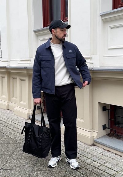 Chaqueta azul marino con dos bolsillos delanteros, sudadera blanca, pantalones negros, bolso tote negro, y zapatos deportivos plateados con gorra negra.