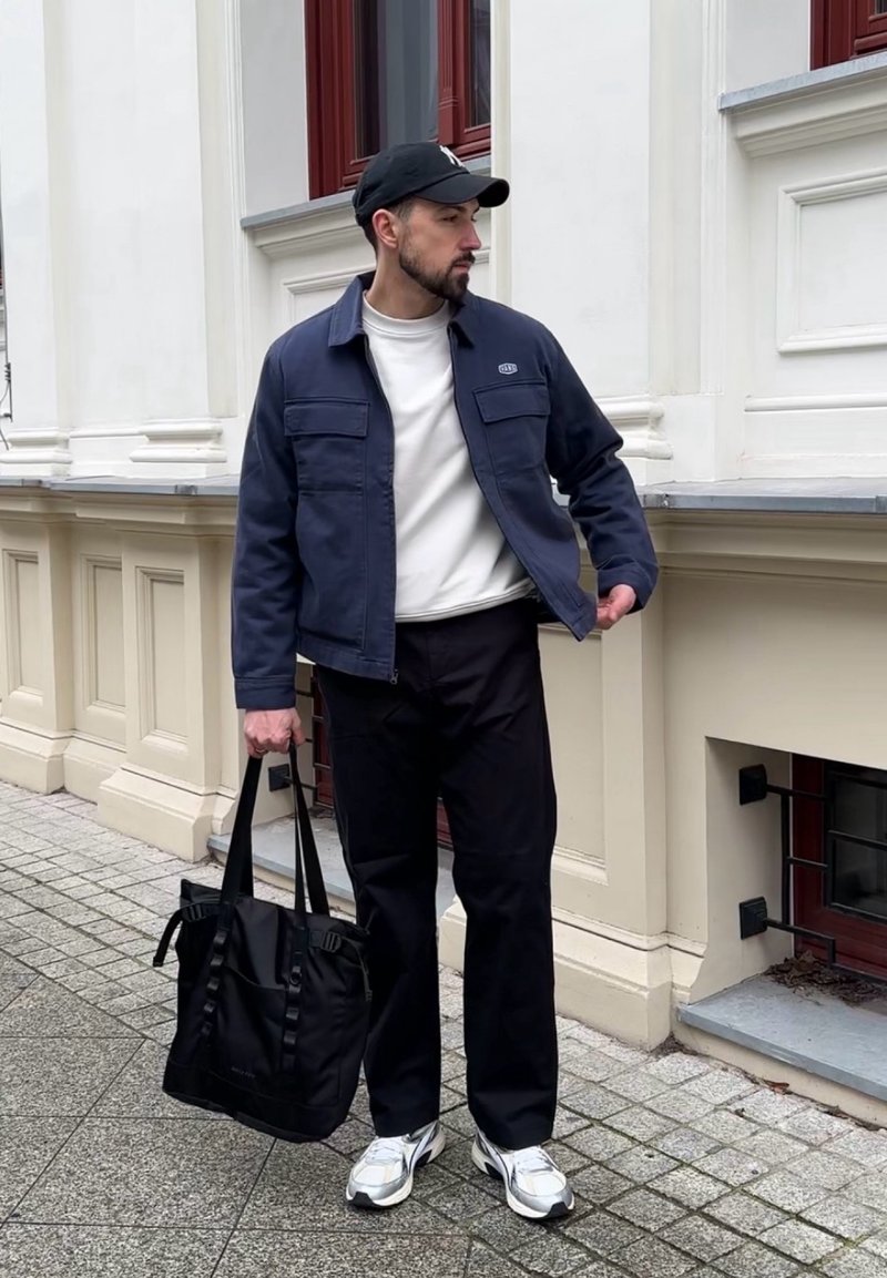 Veste bleue marine avec deux poches frontales, sweat-shirt blanc, pantalon noir, portant un sac fourre-tout noir, et chaussant des chaussures de sport argentées ainsi qu'une casquette noire.