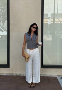 Grå ærmeløs striktop med forknapper, hvide baggy bukser, sorte flip-flops og en tekstureret beige håndtaske. Holder en kaffekop.