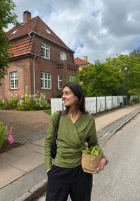 Zielona lniana bluzka z luźnymi czarnymi spodniami, trzymająca brązową torbę papierową z zielonymi warzywami. W tle znajduje się czerwona ceglana willa oraz zieleń.