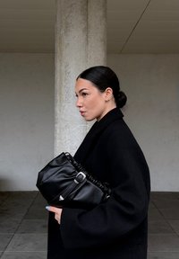 Sac en cuir noir à design froncé avec ferrures argentées, tenu contre un manteau sombre. En arrière-plan, un mur en béton clair.