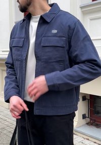 Veste en toile bleu marine avec un col à revers, deux poches avant et un petit logo brodé. Présente une coupe décontractée et un tissu texturé.