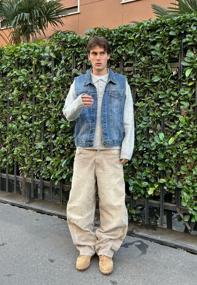 Chaleco de denim sobre un suéter de lana gris claro, pantalones de pana color beige, y botas beige. De pie frente a un seto verde.