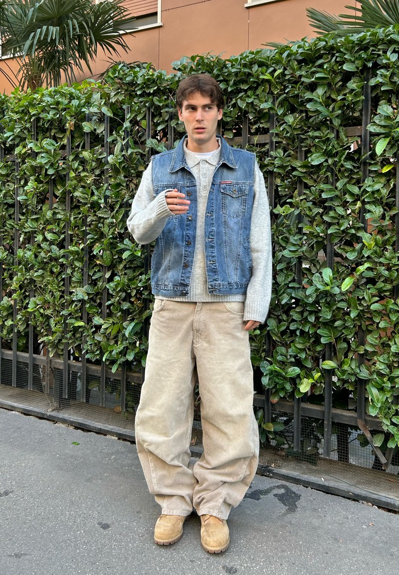 Jeansweste über einem hellgrauen Strickpullover, braunen Cordhosen und beigen Stiefeln. Steht vor einer grünen Hecke.