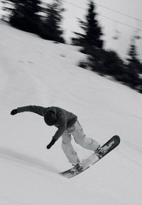 Person som snowboarder i et snøfylt bakke, iført en mørk jakke og lyse bukser, med hjelm og hansker, som utfører et triks.