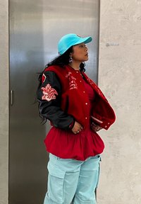 Punane ja must varsity jakk broderieerimisega, kantud punase pluusi ja volüümi andva punase seeliku kohal, koos helesiniste cargo-pükstega ja sinise mütsiga.