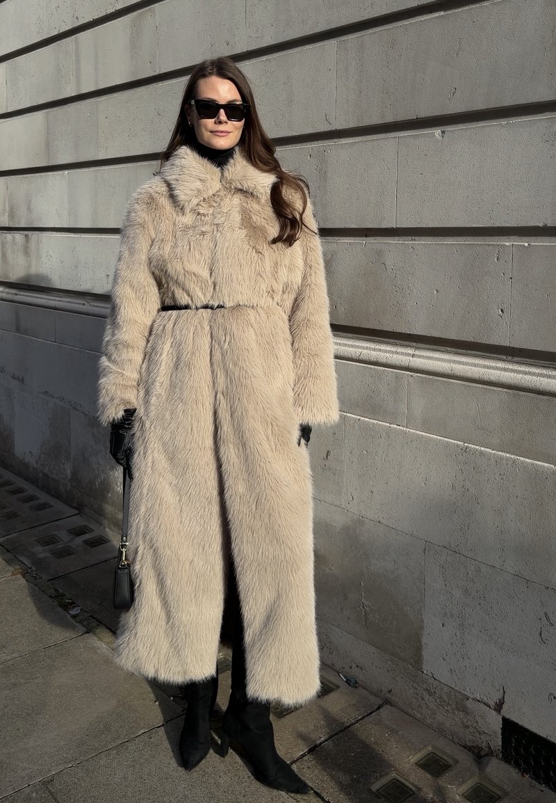 Manteau en fausse fourrure beige avec un col montant, une taille ceinturée, des manches longues et des poches latérales. Porté sur un col roulé noir et un pantalon ajusté.