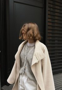 Personne aux cheveux courts et ondulés portant un manteau beige, un cardigan gris et un pantalon blanc, debout devant des panneaux verticaux sombres et des volets.